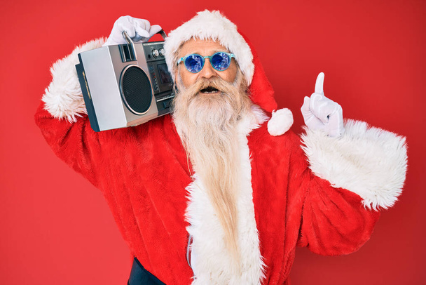 Old senior man wearing santa claus costume and boombox surprised with an idea or question pointing finger with happy face, number one  - Foto, afbeelding