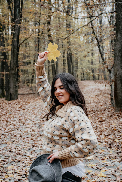 A vertical shot of a beautiful Caucasian woman holding a maple leaf over her head and having a walk in the woods during autumn - Photo, image