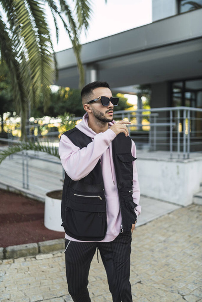 A vertical portrait of an attactive elegant man in a sporty outfit and stylish sunglasses posing against a modern building - Foto, Bild