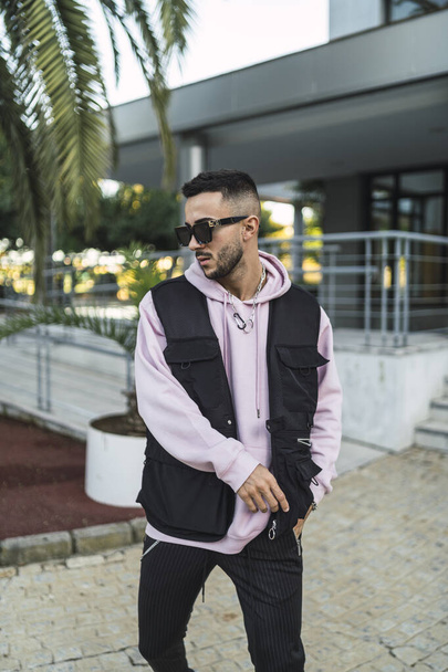 A vertical portrait of an attactive elegant man in a sporty outfit and stylish sunglasses posing against a modern building - Zdjęcie, obraz