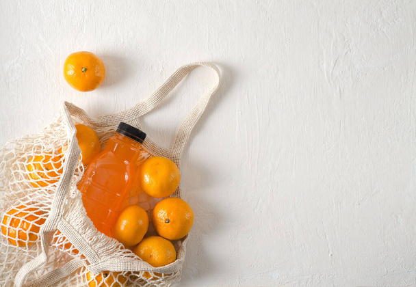 Getränk aus Mandarinen in der Flasche und Mandarinenfrüchte liegen in einem Netzbeutel auf hellem Hintergrund mit Kopierraum, selektiver Fokus, horizontaler Hintergrund - Foto, Bild