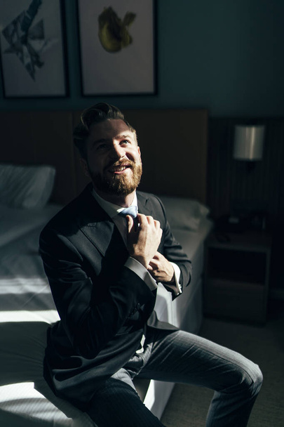 A shallow focus of a cheerful adult male with a beard wearing an elegant suit and sitting on a bed - Valokuva, kuva