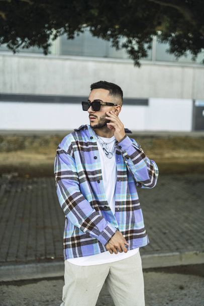 A vertical portrait of a handsome fashionable man in a check shirt and stylish sunglasses posing outdoors - Foto, afbeelding