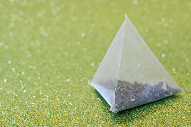 Una bolsa de té piramidal con té verde, rodajas de limón y menta se encuentra en una superficie dorada brillante. Un trago para hígados largos. Macro, bokeh. Profundidad superficial del campo.  - Foto, Imagen