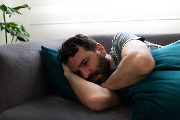 Sad and depressed hispanic man in his 30s lying on his side on the sofa and hugging a cushion - Photo, Image
