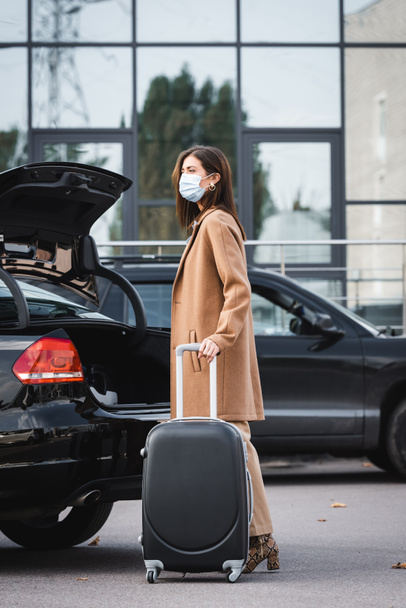 стильная женщина в осеннем наряде и медицинской маске смотрит в сторону, стоя рядом с открытым багажником автомобиля с чемоданом - Фото, изображение