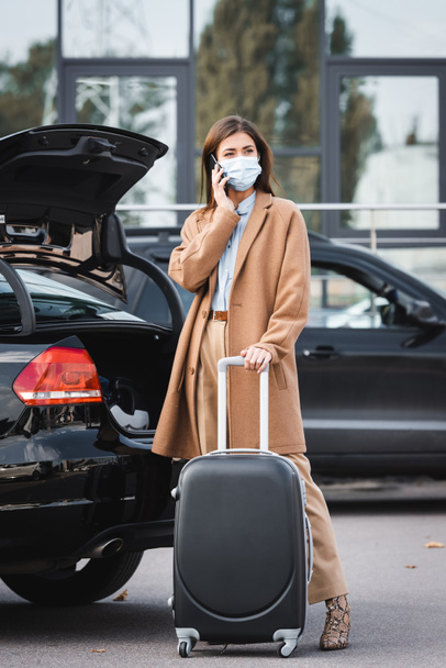 стильная женщина в плаще разговаривает по смартфону, стоя с чемоданом возле багажника - Фото, изображение