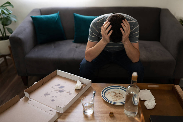Triste hombre adulto sentado en la sala de estar y sosteniendo la cabeza con las manos delante de una mesa con una caja vacía de pizza y alcohol - Foto, Imagen