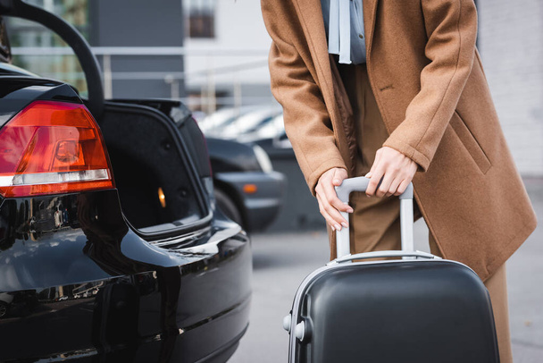 partial view of woman in autumn clothes holding suitcase near open car trunk - Foto, Bild