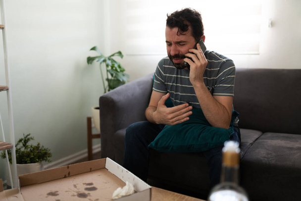 Unglücklicher erwachsener Mann telefoniert und sitzt auf dem Sofa vor einem Tisch mit einer leeren Schachtel Pizza und Alkohol - Foto, Bild