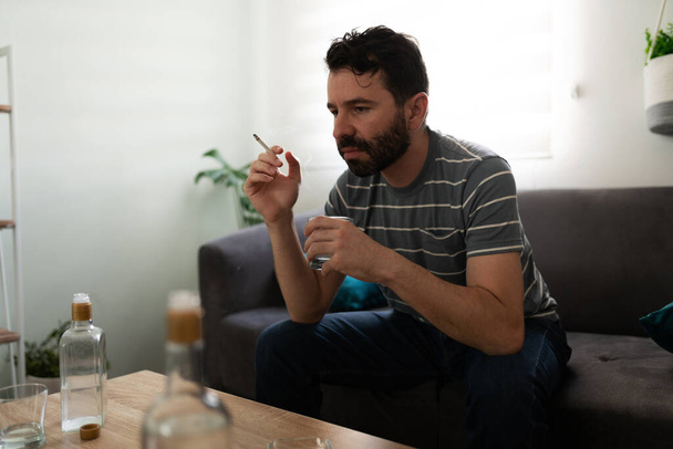 Vue latérale d'un homme alcoolique triste fumant une cigarrete et buvant de la vodka, assis seul dans son canapé - Photo, image