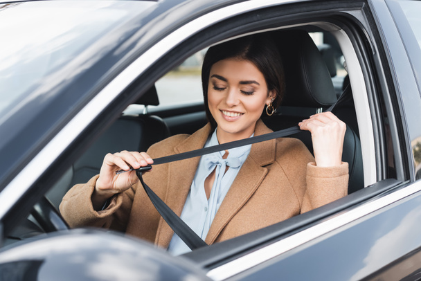 mujer con estilo en traje de otoño ponerse el cinturón de seguridad mientras está sentado en el coche en primer plano borrosa - Foto, Imagen