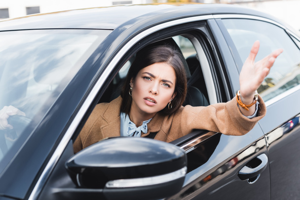 niezadowolona kobieta wyglądająca przez okno boczne i wskazująca ręką podczas prowadzenia samochodu na niewyraźnym pierwszym planie - Zdjęcie, obraz