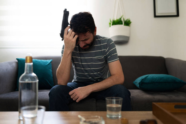 Homme adulte alcoolique et déprimé pleurant en pensant à se suicider avec une arme près de sa tête, assis sur sa maison devant une bouteille d'alcool  - Photo, image