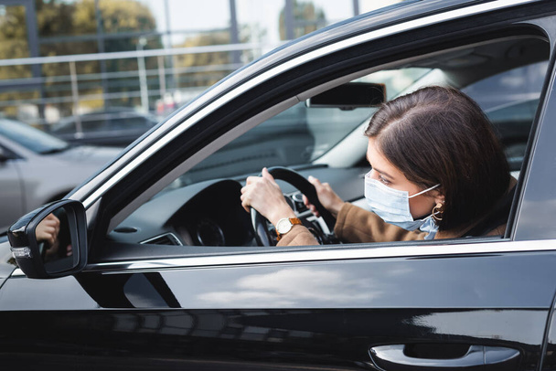 молодая женщина в медицинской маске смотрит из бокового окна во время руля автомобиля - Фото, изображение