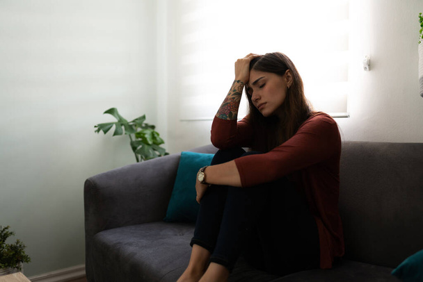 Nuori 20-vuotias nainen näyttää surulliselta ja surumieliseltä. Latinalainen nainen istuu kotona sohvalla ja halaa polviaan - Valokuva, kuva