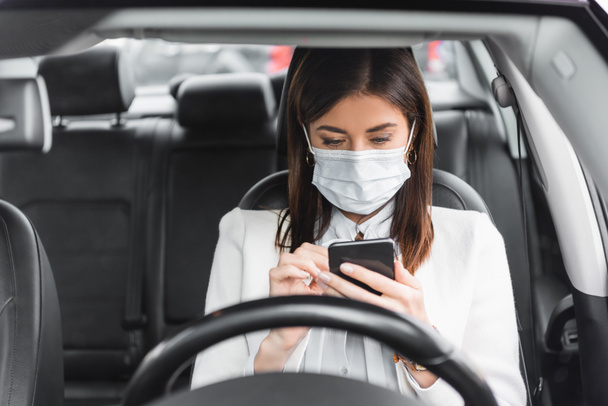 молодая женщина в медицинской маске отправляет сообщения на смартфоне в машине на размытом переднем плане - Фото, изображение