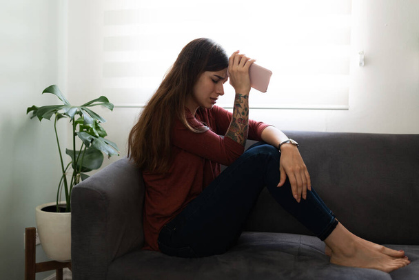 Vista lateral de una triste y deprimida joven sentada en el sofá con su teléfono celular. Mujer latina que parece solitaria y melancólica en su casa - Foto, Imagen