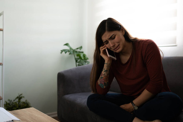 Aantrekkelijke Spaanse jonge vrouw huilend en pratend aan de telefoon over haar problemen. Depressieve en eenzame vrouw in de woonkamer - Foto, afbeelding