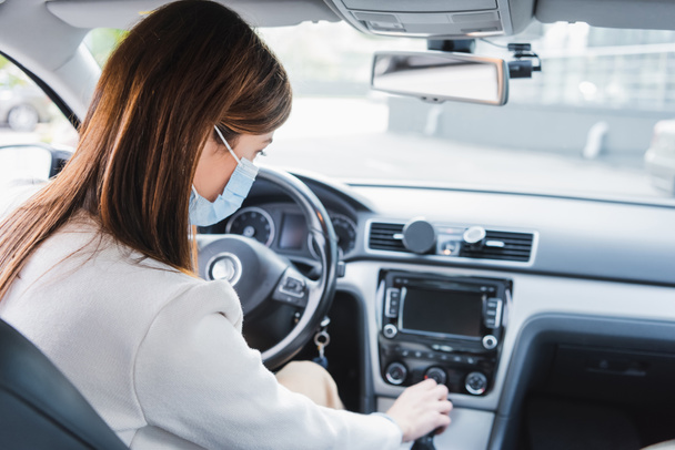 młoda kobieta w masce medycznej przesuwa dźwignię skrzyni biegów siedząc w samochodzie - Zdjęcie, obraz