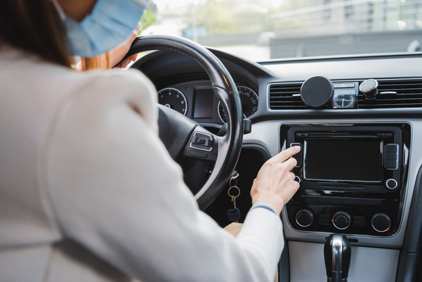 przycięty widok kobiety w masce medycznej naciskając przycisk na desce rozdzielczej podczas siedzenia w samochodzie - Zdjęcie, obraz