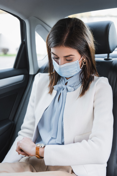 stylowa kobieta w masce medycznej patrząc na zegarek podczas jazdy samochodem na tylnym siedzeniu - Zdjęcie, obraz