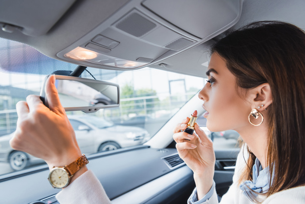 стильна жінка тримає помаду, дивлячись в дзеркало заднього виду автомобіля
 - Фото, зображення