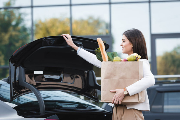 smiling woman opening car trunk while holding shopping bag with food - Фото, изображение