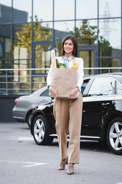 mujer joven con estilo sosteniendo bolsa de compras con comida mientras camina a lo largo de aparcamiento - Foto, imagen