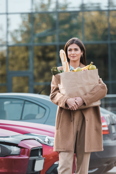 young woman in trench coat carrying shopping bag with food while walking along car parking - Фото, изображение