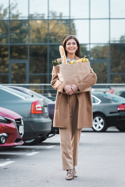 Sonbahar kıyafetli neşeli bir kadın alışveriş çantası ve yiyecek taşırken araba parkında yürüyor. - Fotoğraf, Görsel