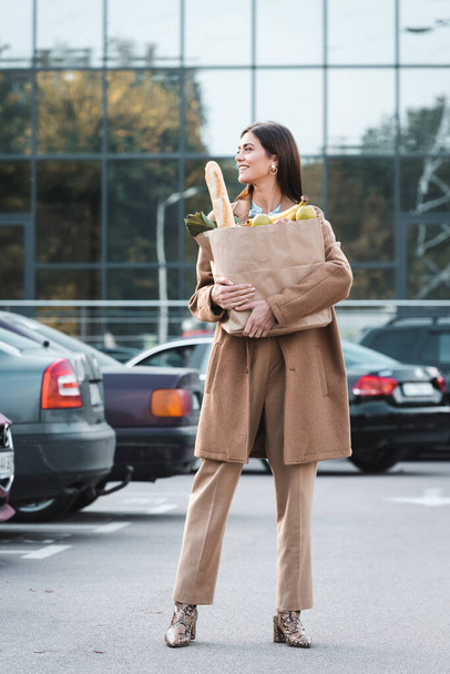 веселая женщина в стильном осеннем наряде смотрит в сторону, держа сумку с едой - Фото, изображение