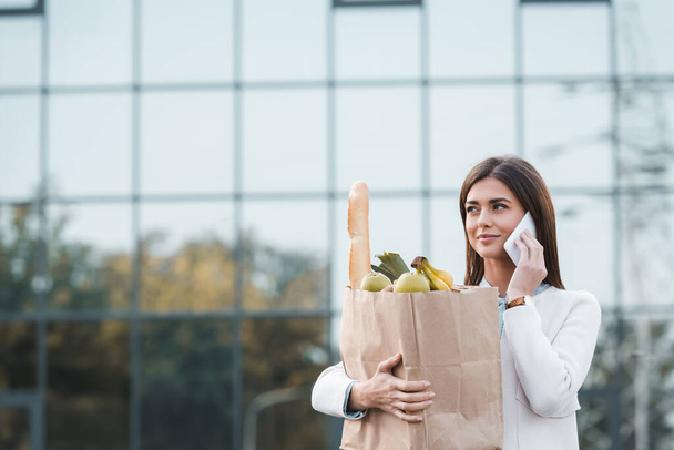 positive young woman looking away while holding shopping bag with food and talking on smartphone - Foto, Bild