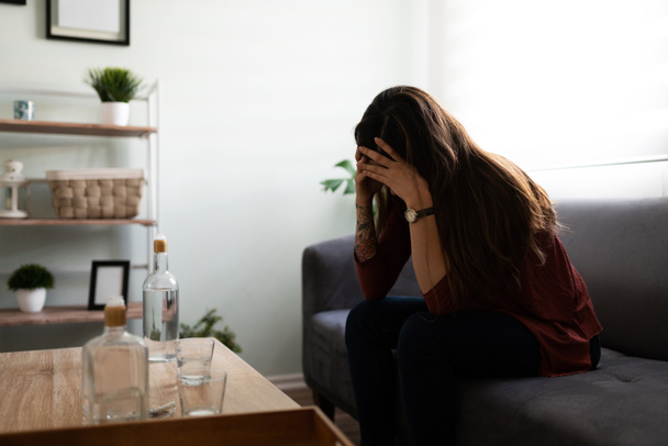 Side view of an alcoholic young woman sitting alone on the sofa looking stressed and sad. Depressed woman sitting holding her head  - Photo, Image