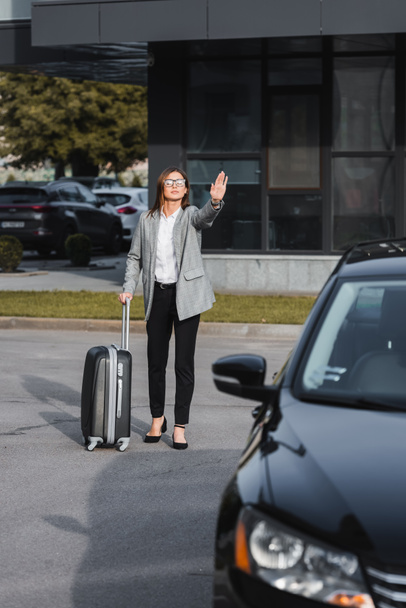 κομψή επιχειρηματίας με βαλίτσα κουνώντας το χέρι κοντά στο μαύρο αυτοκίνητο σε θολή πρώτο πλάνο - Φωτογραφία, εικόνα