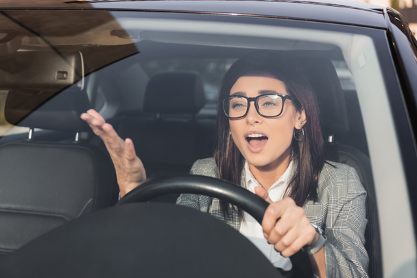 сердитая женщина кричит и жестикулирует во время вождения автомобиля на размытом переднем плане - Фото, изображение