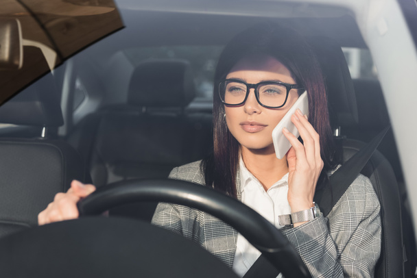 femme d'affaires élégante dans les lunettes de direction de voiture tout en parlant sur téléphone mobile au premier plan flou - Photo, image