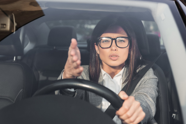 Hoşnutsuz kadın kameraya bakıyor ve ön planda bulanık bir şekilde araba kullanırken elini işaret ediyor. - Fotoğraf, Görsel
