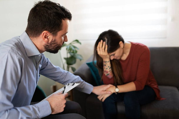 Hopeless young woman looking sad while holding her head during therapy. Male psychiatrist giving comfort to a depressed female patient  - Photo, Image