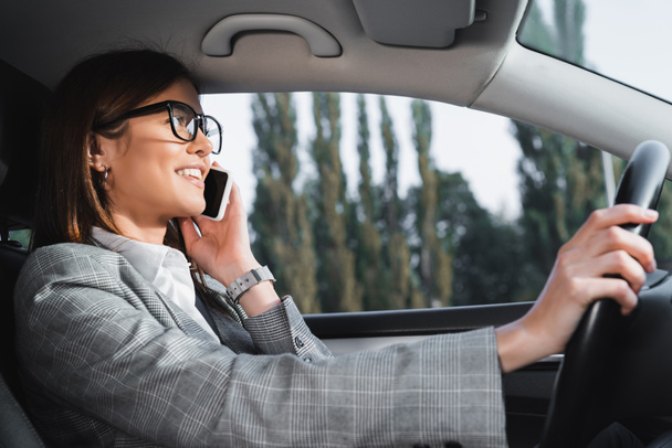 весела бізнес-леді в окулярах за кермом автомобіля і розмовами на смартфоні
 - Фото, зображення