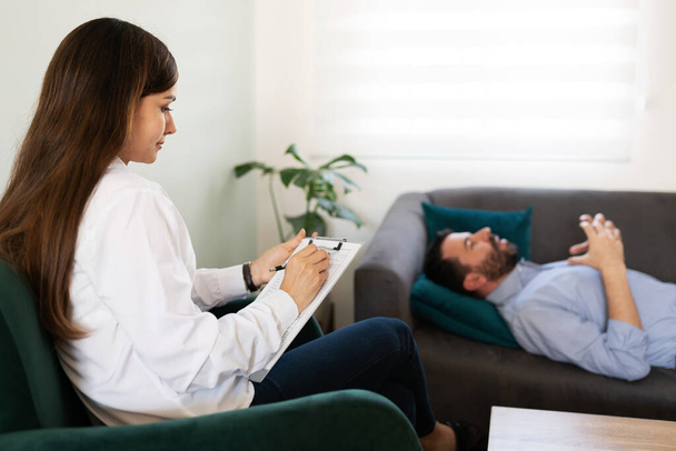 Jovem pshycologist feminino escrevendo em seu gráfico durante uma sessão de terapia com seu paciente do sexo masculino  - Foto, Imagem