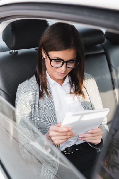 happy businesswoman in eyeglasses using digital tablet while riding in car on blurred foreground - Photo, Image