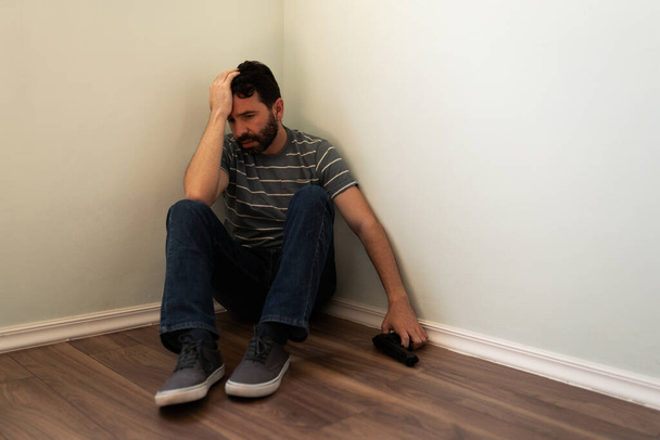 Latinský dospělý muž s depresí problém sedí v rohu s pistolí a přemýšlí o sebevraždě  - Fotografie, Obrázek