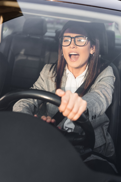 сердитая женщина кричит и пищит во время вождения автомобиля и смотрит вперед на размытый передний план - Фото, изображение