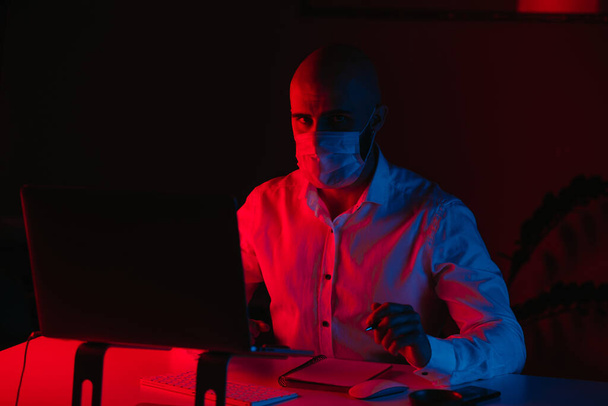 Ein glatzköpfiger Mann mit medizinischer Gesichtsmaske arbeitet zu Hause an einem Laptop. Ein Kerl starrt, während er Notizen macht. Ein männlicher Mitarbeiter mit einem Stift vor dem Computer unter blauem und rotem Licht. - Foto, Bild