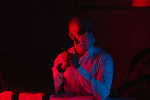 Ein glatzköpfiger Mann mit medizinischer Gesichtsmaske arbeitet zu Hause an einem Laptop. Ein Typ starrt weg. Ein männlicher Angestellter mit verschränkten Armen vor dem Computer unter blauem und rotem Licht. - Foto, Bild