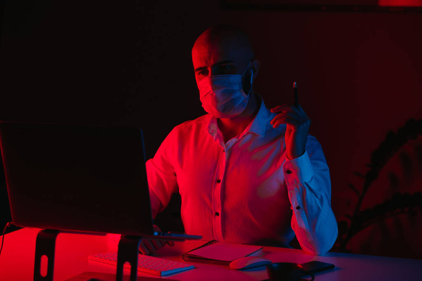 A bald man in a medical face mask is working remotely on a laptop at home. A guy is reading and doing notes. A male employee with his arms folded in front of the computer under blue and red light. - Valokuva, kuva