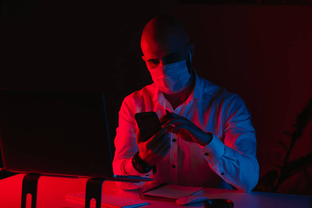 Een kale man met een medisch gezichtsmasker werkt thuis op afstand op een laptop. Een man leest het nieuws op een smartphone. Een mannelijke werknemer met een pen voor de computer onder blauw en rood licht. - Foto, afbeelding