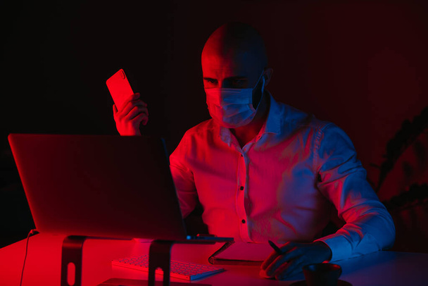 Ένας φαλακρός με μάσκα ιατρικού προσώπου δουλεύει εξ αποστάσεως σε ένα λάπτοπ στο σπίτι. Ένας τύπος με ακουστικά κρατάει ένα κινητό. Ένας άντρας υπάλληλος με στυλό μπροστά στον υπολογιστή κάτω από μπλε και κόκκινο φως.. - Φωτογραφία, εικόνα