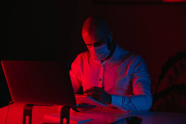 Ένας φαλακρός με μάσκα ιατρικού προσώπου δουλεύει εξ αποστάσεως σε ένα λάπτοπ στο σπίτι. Ένας τύπος με ακουστικά κρατάει σημειώσεις. Ένας άντρας υπάλληλος με στυλό μπροστά στον υπολογιστή κάτω από μπλε και κόκκινο φως.. - Φωτογραφία, εικόνα